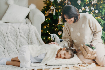 Obraz na płótnie Canvas Attractive brunette smiling woman stands on knees with present boxes, little daughter sleeps near on pillow, has pleasant dreams, enjoy holiday. Winter time, New Year and Christmas. Rest concept