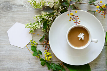 Flatlay with an coffee cup on a saucer with mockup an empty white card framed by branches of green leaves, white lilac flowers and yellow small flowers