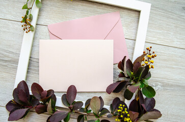Close-up flatlay with white wooden frame and mockup and copyspace pastel pink envelopes and blank postcard framed by branches of burgundy and green leaves, white lilac flowers and yellow small flowers