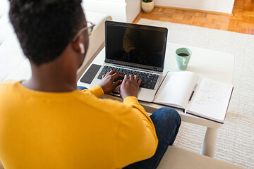 Handsome African American man working on laptop computer while sitting behind desk in cozy living room. Freelancer working from home. Browsing internet, using social network.