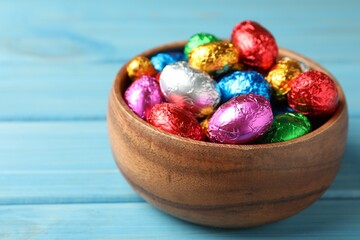 Bowl with chocolate eggs wrapped in colorful foil on light blue wooden table, closeup