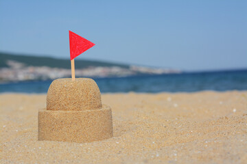 Fototapeta na wymiar Beautiful sand castle with red flag on beach near sea, space for text