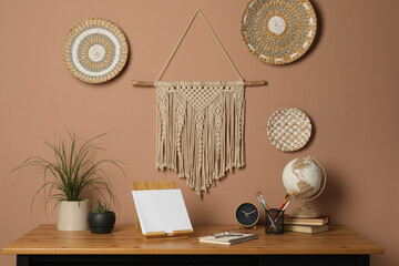 Stylish macrame and wicker wall decor hanging above wooden table with houseplants and stationery
