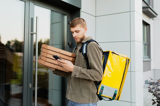 A courier with a mobile phone checks the apartment number standing at the entrance. A man holds pizza boxes