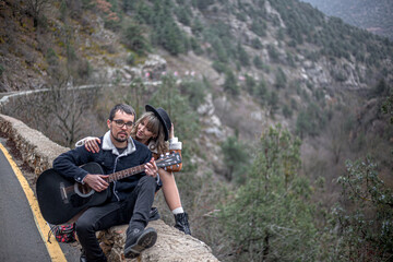 Naklejka na ściany i meble back view of couple in love walking in the countryside road against the mountain and road, man holding the guitar, people hitchhiking . A man gently hugs a woman