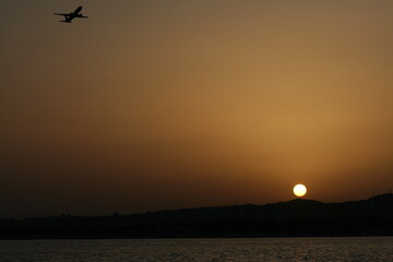 Foto del tramonto del sole dietro le colline viste dal mare, con un aereo in fase di decollo