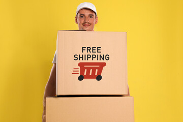 Happy young courier with cardboard boxes on yellow background. Free shipping
