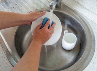 Women's hands wash white dishes in a metal sink with a blue sponge. White ceramic plate and cup. Water is flowing from the kitchen faucet.