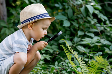 Summer holidays. A small child in a hat looks at the leaves with a magnifying glass. A preschool boy studies nature with a magnifying glass.  Education, excursions, research concept.