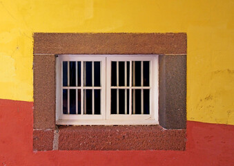 a white closed wooden house window in a stone frame on a brightly painted red and yellow wall in typical portuguese colors