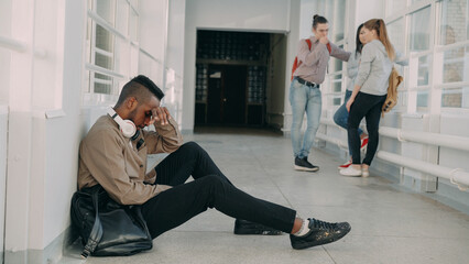 Fototapeta na wymiar View of upset serious african-american male student sitting on floor in white spacious university corridorwhile his groupmates are standing communicating.