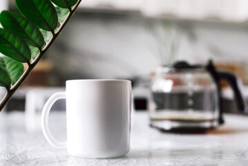 Mock-up of a white mug with coffee or tea is on the table with a glass teapot.