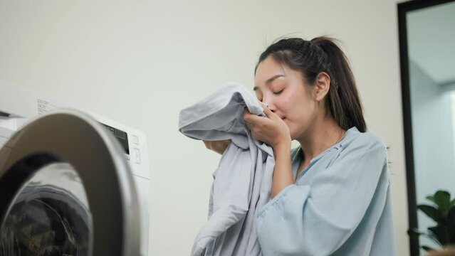 Happy housewife opens the front of the washing machine and smiles to receive the laundry that has been washed at house. Woman smelling fragrant clean clothes washed from softener and washing machine.