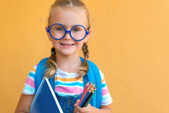 portrait smiling school girl in glasses with school backpack holds colored pencils and blue notebook. ready for school. back to school. isolated on a yellow background copy space. advertising sale