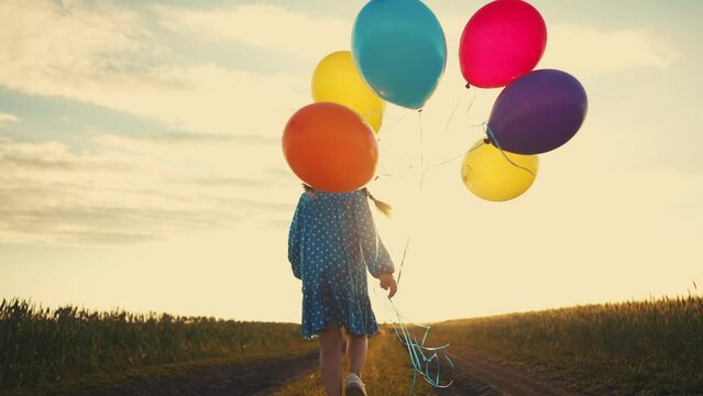 little girl runs with balloons in the park in nature. happy family holiday birthday kid dream concept. a little sun girl with colorful balloons runs with her back along the road in the field park