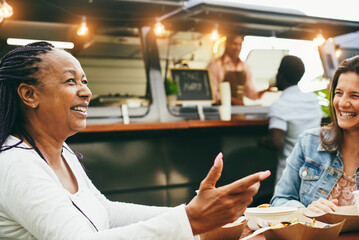 Happy multiracial senior women eating at food truck restaurant outdoor - Focus on african female face