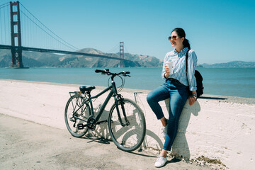 stylish asian woman leaning on short white wall is having a coffee break from bike riding near...