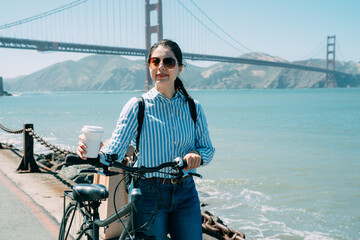 cheerful asian girl carrying bike and walking with coffee is enjoying the scenery at marina with...