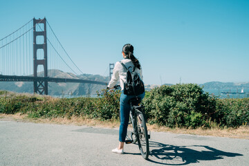 rear view asian female backpacker is admiring great golden gate bridge in the distance on bike on a...