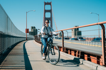 full length shot carefree asian girl wearing sunglasses is riding a bicycle on golden gate bridge...