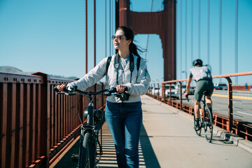 Asian woman is looking into distance and enjoying the view while walking with a bike on golden gate bridge at leisure on a sunny day in San Francisco California - Powered by Adobe