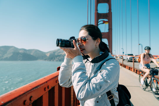 portrait of asian female professional photographer is taking photos with a digital camera on golden gate bridge in San Francisco California USA in the sun.