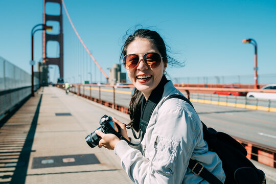 cheerful asian woman photographer is turning to smile at the camera while taking pictures on golden gate bridge in San Francisco California on a sunny day.