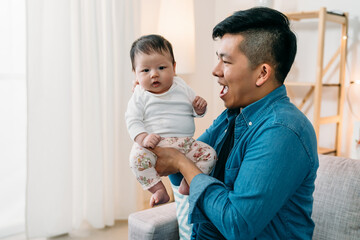 asian cute newborn infant girl is sitting in her father’s hands with innocence while her father is looking and playing with her in a bright living room at home.