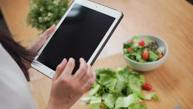 Beautiful woman using tablet to calculate calories and nutrition vegetable salad in weight loss diet. Young female happily cooks a healthy breakfast in kitchen in the morning. Diet food concept.