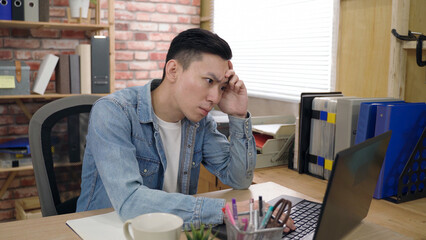 asian young male is propping his head and staring at the monitor while pondering on a difficult...