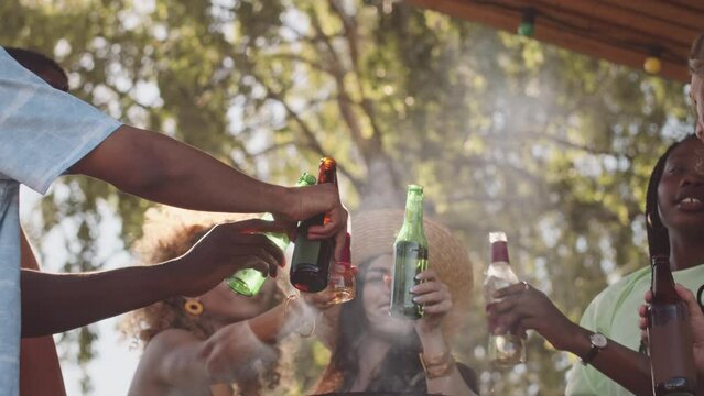Low angle slowmo of joyful young multiethnic friends having fun at barbecue party in summerhouse, clinking beer bottles and chatting