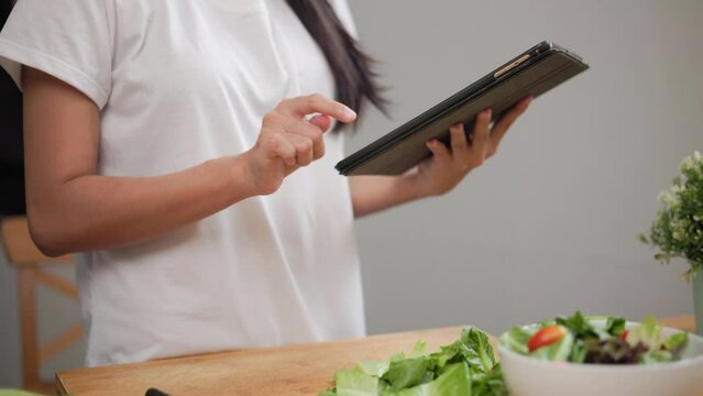 Beautiful woman using tablet to calculate calories and nutrition vegetable salad in weight loss diet. Young female happily cooks a healthy breakfast in kitchen in the morning. Diet food concept.