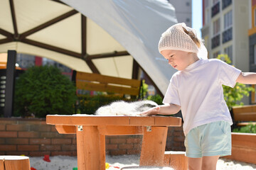 Modern playground. A cute girl shakes sand off a wooden children's table. Outdoor activities. Summer entertainment