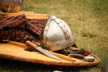 Equipment, costume and props of a Viking reenator lying on the ground outdoors; helmet, wooden handled knife, sword, shield and tunic. - obrazy, fototapety, plakaty