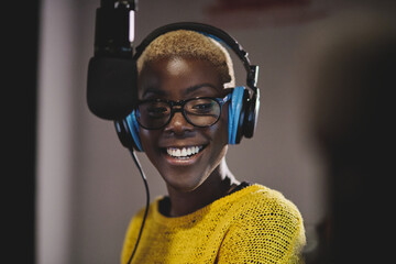 Optimistic ethnic female radio host in bright yellow sweater headphones and glasses recording...