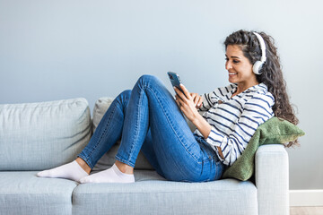 Woman in headphones holding smart phone and gesturing while sitting on the couch at home. Photo of...