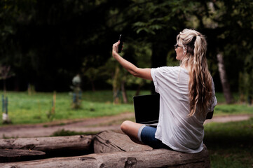 A girl with a laptop works while walking in the park in a cozy place, enjoys it. Online learning, freelancing and fun concept, talk by mobile cell phone look aside. People urban lifestyle concept