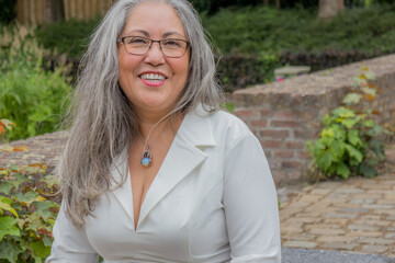 Portrait of mature woman with happy smiling expression, attentively looking at camera, straight gray hair, tanned skin, glasses, light makeup, relaxed day in garden, green vegetation in background