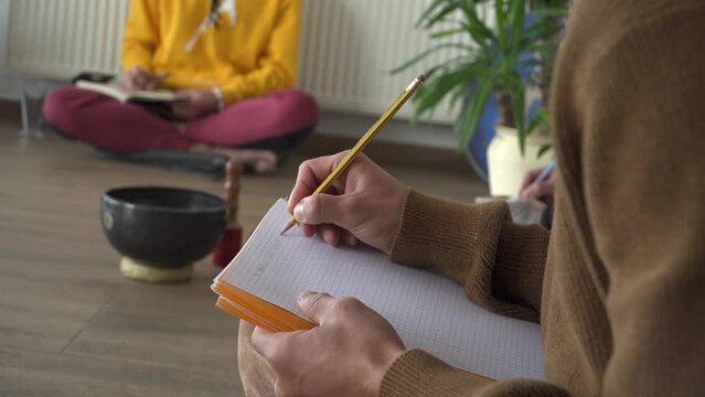 Close up view of person in brown sweater writing on lined notebook paper with pencil. Journaling, recording thoughts, feelings and experiences in a diary. People taking notes seated on floor.