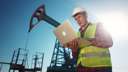 oil industry. engineer next to an oil rig. oil production business finance concept. gas production. a worker with a laptop monitors oil production next to an station. a worker sun in hard hat works