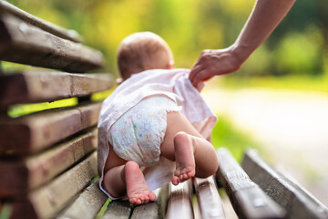 Baby in diaper crawling on a park bench on a sunny summer day. Mother's hand helps and protects the child. Happy kid learn and play. - obrazy, fototapety, plakaty