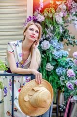 Stunning portrait of a blond girl in an elegant dress with a straw hat near a city cafe decorated with flowers on a summer day. Fashion and beauty concept.
