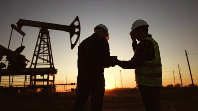 oil production. two workers a work next to an oil pump at sunset silhouette. industry business oil production concept. lifestyle engineers in hardhats studying oil production figures