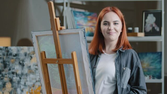 Cheerful carefree Caucasian woman artist girl painter with red hair draws in art studio creates picture listens to music dances has fun looking at camera funny laughing enjoys hobby drawing dancing