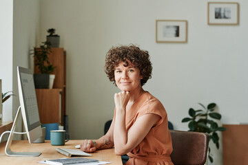Portrait of young businesswoman sitting at her workplace in front of computer monitor at office and...