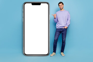 Full size photo of nice young brunet guy stand index big telephone wear sweater jeans footwear isolated on blue background