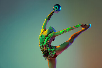 Portrait of young girl, rhythmic gymnast athlete training with ball isolated over multicolored studio background in neon light