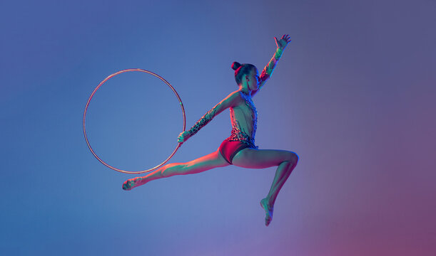 Portrait of young sportive girl, female rhythmic gymnast training, performing with hoop isolated on blue purple background in neon light