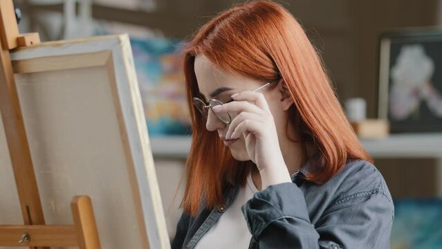 Caucasian woman artist girl painter with red hair put on eyeglasses wearing glasses start art work with oil acrylic paints creates picture drawing painting on canvas inspired student learning draw