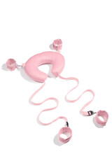 Detailed shot of a pink velvety neck pillow with clasps and buckles on belts connected with pink handcuffs and shackles. The accessory for erotic games is located on the white backdrop.